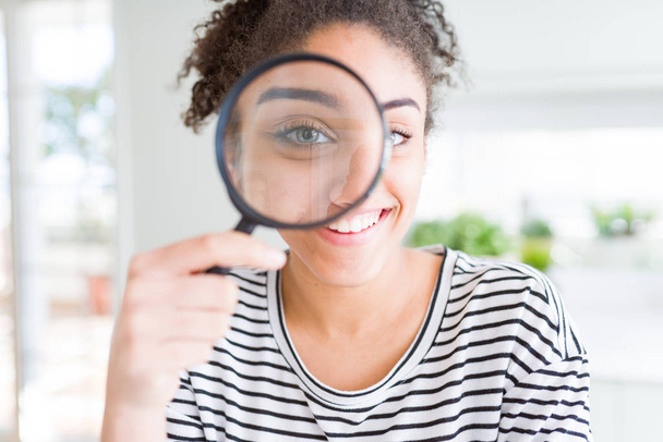 Young african american woman looking through magnifying glass with a happy face standing and smiling with a confident smile showing teeth - Foto, Imagem