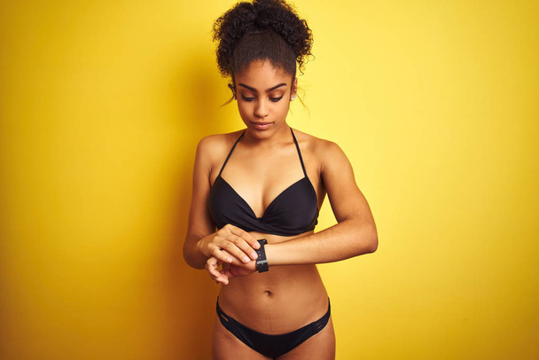 Afrika Amerikan kadın tatil üzerinde izole sarı arka plan üzerinde ayakta giyen bikini bilek saatindeki saati kontrol etme, rahat ve emin - Fotoğraf, Görsel