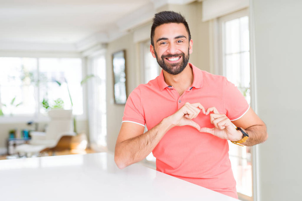 Knappe Spaanse man thuis met het dragen van casual t-shirt glimlachend in liefde met hartsymbool en vorm met de handen. Romantische concept. - Foto, afbeelding