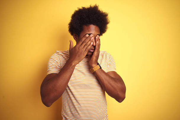 amerikanischer Mann mit Afro-Haaren und gestreiftem T-Shirt, der vor isoliertem gelben Hintergrund steht, reibt sich die Augen vor Müdigkeit und Kopfschmerzen, schläfriger und müder Ausdruck. Sehschwäche - Foto, Bild