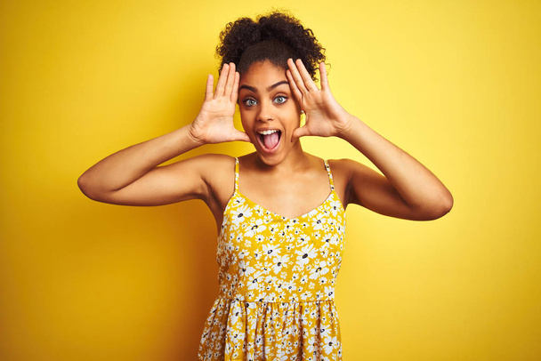African american woman wearing casual floral dress standing over isolated yellow background Smiling cheerful playing peek a boo with hands showing face. Surprised and exited - Foto, afbeelding