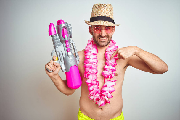 Young man wearing summer hat and hawaiian lei flowers holding water gun over isolated background with surprise face pointing finger to himself - Photo, Image