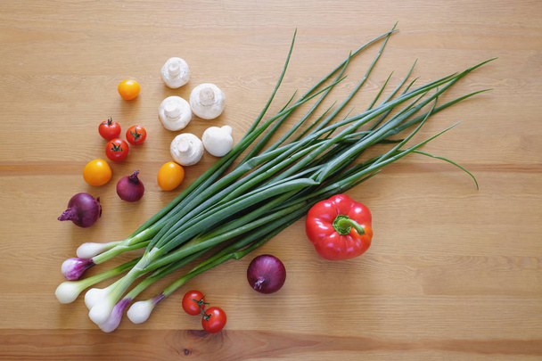 Λαχανικά στο ξύλινο φόντο. Βιολογικά τρόφιμα και φρέσκα λαχανικά. Πράσινα κρεμμύδια, ντοματίνια, κόκκινη πιπεριά, κρεμμύδια και ροδάκια στο τραπέζι. - Φωτογραφία, εικόνα
