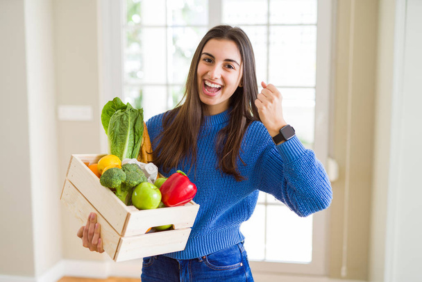 Beautiful young woman holding wooden box full of healthy groceries screaming proud and celebrating victory and success very excited, cheering emotion - Fotoğraf, Görsel