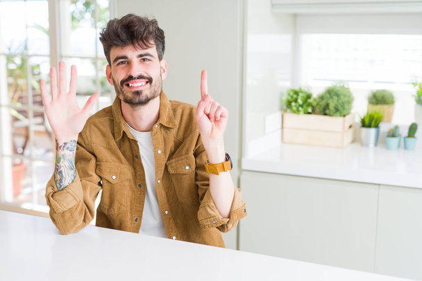 白いテーブルの上に座ってカジュアルなジャケットを着た若い男は、自信を持って幸せに微笑みながら、指番号6を示し、指を上に向けます. - 写真・画像