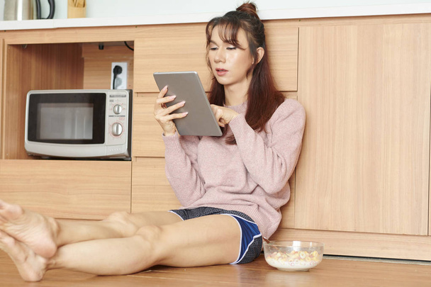 Jovem mulher descansando em casa ela sentada no chão na cozinha e surfar a rede no tablet pc durante o café da manhã
 - Foto, Imagem