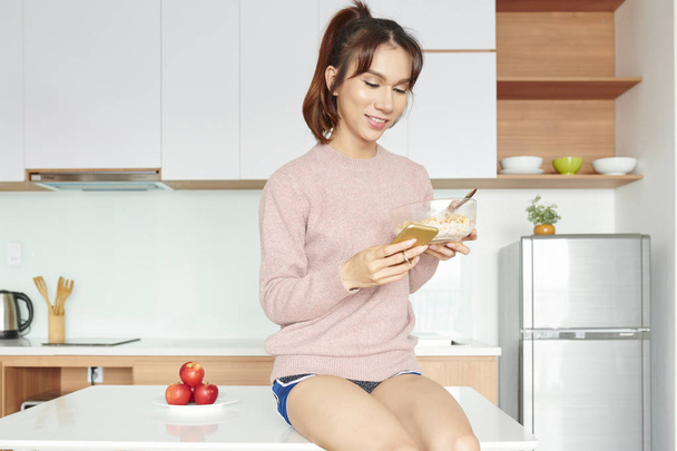 asiatische junge Frau sitzt auf dem Tisch und liest eine Nachricht auf ihrem Handy und lächelt, während sie Cornflakes mit Milch zum Frühstück isst - Foto, Bild