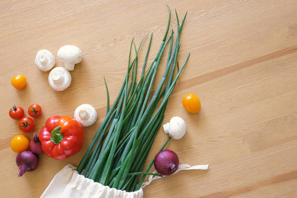 Σετ προϊόντων σε μια τσάντα Eco από βαμβάκι σε ένα τραπέζι, κόκκινο κρεμμύδι, κόκκινο πιπέρι, μανιτάρια και ντοματίνια. Η έννοια της μηδενικής σπατάλης. - Φωτογραφία, εικόνα