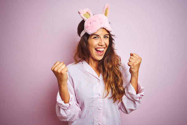 Jonge vrouw het dragen van pyjama en slaap masker staande over roze geïsoleerde achtergrond vieren verrast en verbaasd voor succes met armen verheven en open ogen. Winnaar concept. - Foto, afbeelding
