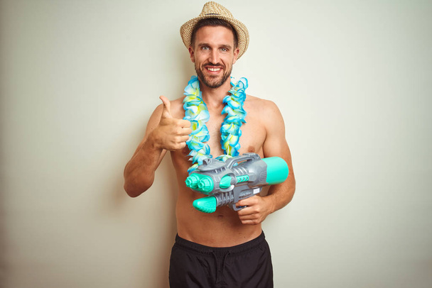 Pohledný muž v havajském lei a vodní pistoli na pozadí šťastný s velkým úsměvem dělá dobře nápis, palec s prsty, výborný nápis - Fotografie, Obrázek