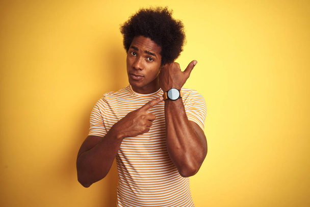 Hombre americano con pelo afro vistiendo camiseta a rayas de pie sobre fondo amarillo aislado En prisa apuntando a ver el tiempo, la impaciencia, mirando a la cámara con expresión relajada
 - Foto, Imagen