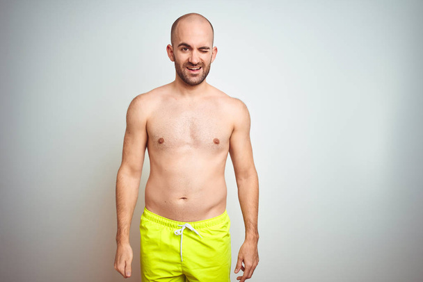 Young shirtless man on vacation wearing yellow swimwear over isolated background winking looking at the camera with sexy expression, cheerful and happy face. - Foto, imagen