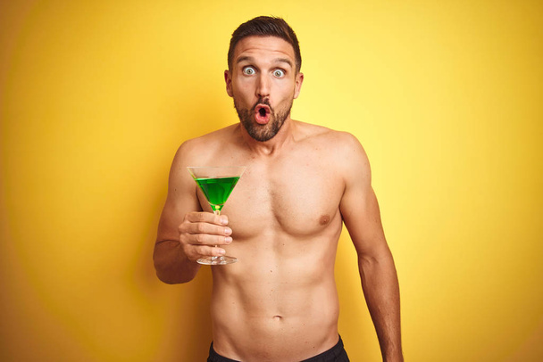 Jeune homme beau torse nu buvant un cocktail d'été sur fond jaune isolé effrayé en état de choc avec un visage surprise, effrayé et excité par l'expression de la peur
 - Photo, image
