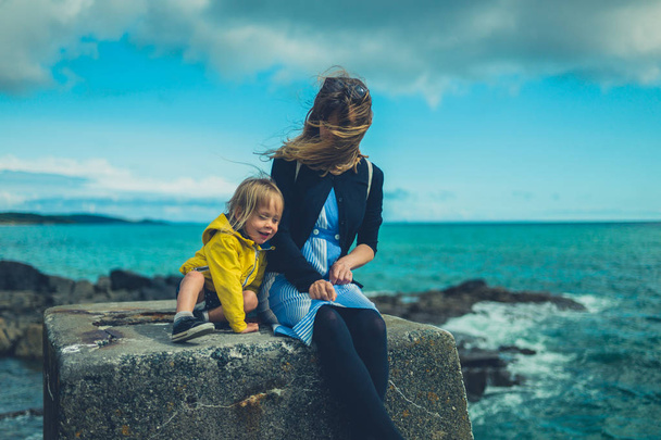 Young mother and toddler sitting on concrete block by sea in summer - Photo, Image