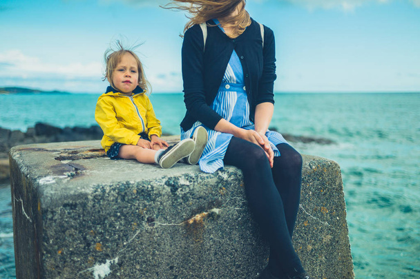 Young mother and toddler sitting on concrete block by sea in summer - Foto, imagen