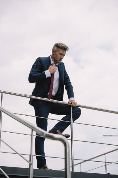 selective focus of upset businessman in suit standing outside and touching tie - Fotoğraf, Görsel