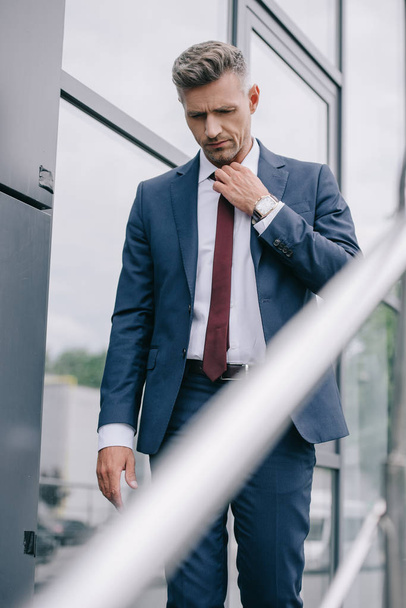 selective focus of frustrated businessman in formal wear touching tie outside near building - Фото, изображение