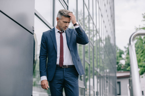 selective focus of upset businessman in formal wear standing near building and touching hair  - Foto, afbeelding