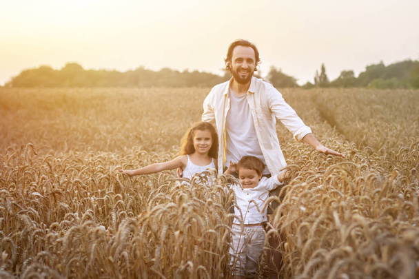 Feliz familia, padre, hijo y niña en sombrero de paja en el campo de trigo al atardecer. El concepto de agricultura ecológica y estilo de vida saludable, alimentación saludable, felicidad y alegría
 - Foto, Imagen