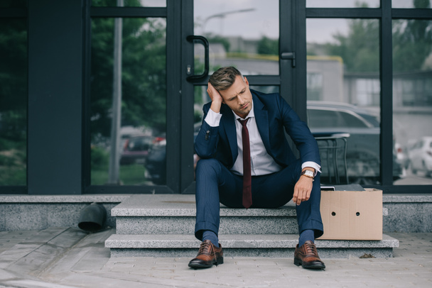 dismissed businessman in suit sitting on stairs near carton box and touching hair  - Photo, Image