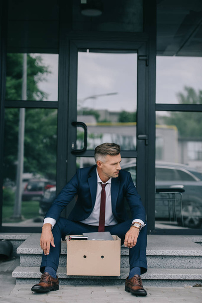 fired businessman in suit sitting on stairs near carton box  - Foto, afbeelding