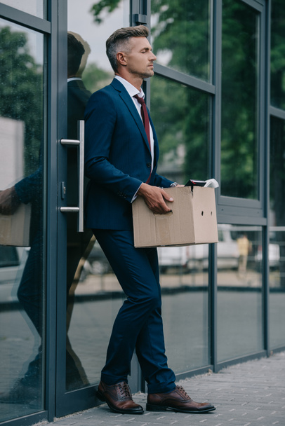 upset and fired businessman standing near building and holding carton box  - Foto, imagen
