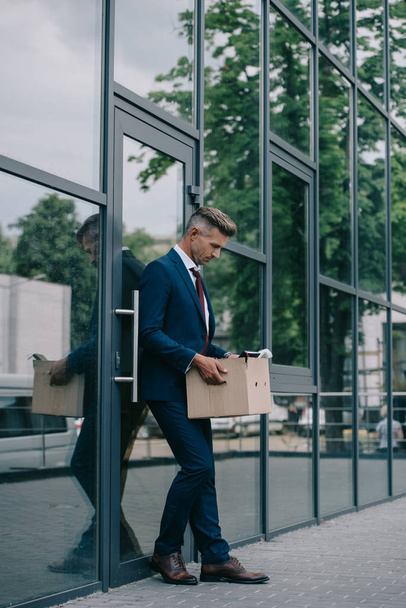upset and fired businessman standing near building and looking at carton box  - Foto, imagen