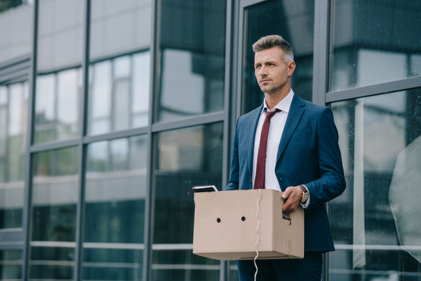звільнений бізнесмен у костюмі, що стоїть біля будівлі з картонною коробкою
  - Фото, зображення