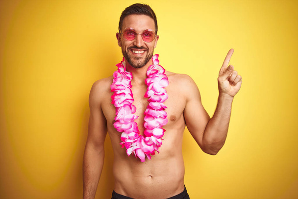 Young handsome shirtless man wearing sunglasses and pink hawaiian lei over yellow background with a big smile on face, pointing with hand and finger to the side looking at the camera. - Zdjęcie, obraz