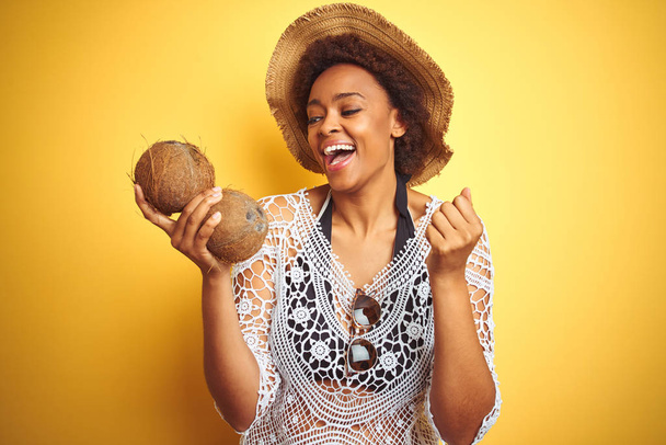 Giovane donna afro americana con i capelli afro tenendo cocco su sfondo giallo isolato urlando orgoglioso e celebrando la vittoria e il successo molto eccitato, emozione tifo
 - Foto, immagini