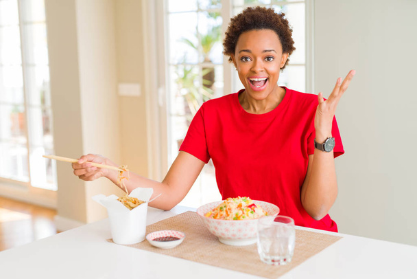 Jeune femme afro-américaine aux cheveux afro mangeant de la nourriture asiatique à la maison très heureuse et excitée, expression gagnante célébrant la victoire en criant avec un grand sourire et les mains levées
 - Photo, image