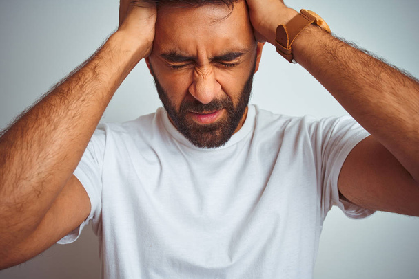 Hombre indio joven con camiseta de pie sobre un fondo blanco aislado que sufre de dolor de cabeza desesperado y estresado debido al dolor y la migraña. Manos en la cabeza
. - Foto, imagen