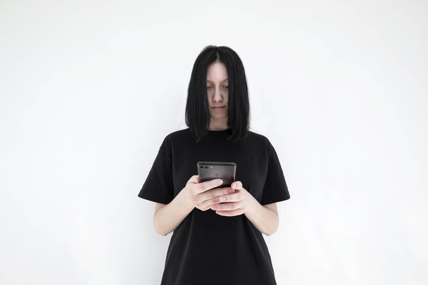 A menina em um vestido preto e com cabelo preto com um smartphone em suas mãos em um fundo branco. A menina está assistindo a um vídeo em um telefone celular
  - Foto, Imagem