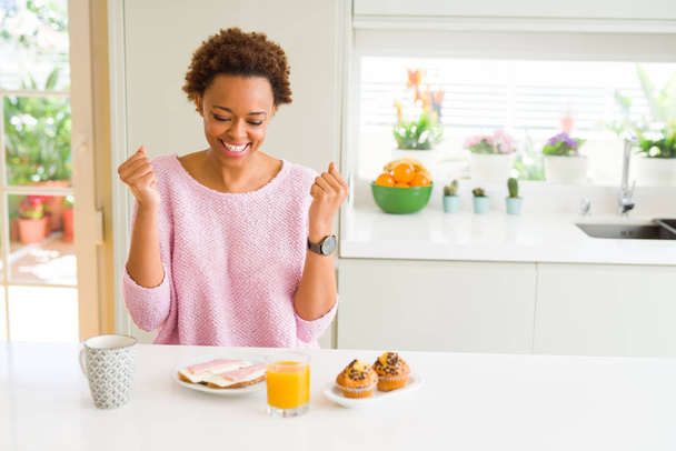 若いアフリカ系アメリカ人女性は、自宅で朝に休憩を食べ、腕を上げて目を開けて成功に驚き、驚きました。勝者のコンセプト. - 写真・画像