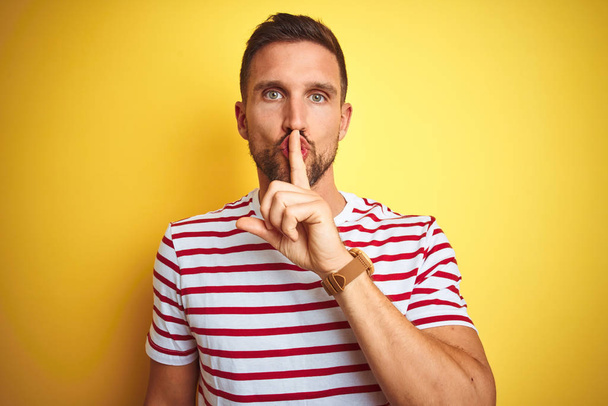 Young handsome man wearing casual red striped t-shirt over yellow isolated background asking to be quiet with finger on lips. Silence and secret concept. - Foto, Imagem