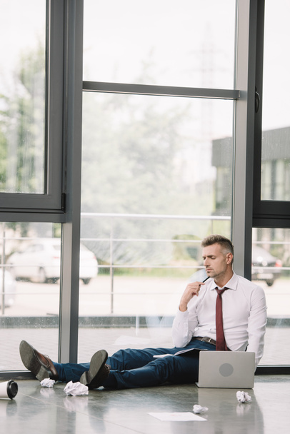 pensive man holding pen while sitting on floor near laptop  - Φωτογραφία, εικόνα