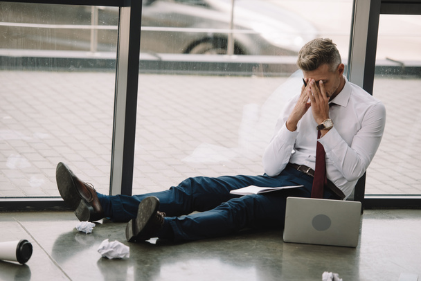 boos zakenman zittend op de vloer en bedekt gezicht met handen in de buurt van laptop - Foto, afbeelding
