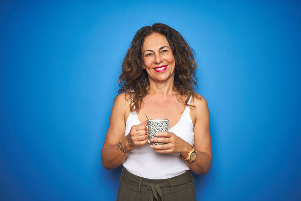 Mujer mayor de mediana edad bebiendo una taza de café sobre fondo azul aislado con una cara feliz de pie y sonriendo con una sonrisa confiada mostrando los dientes
 - Foto, imagen