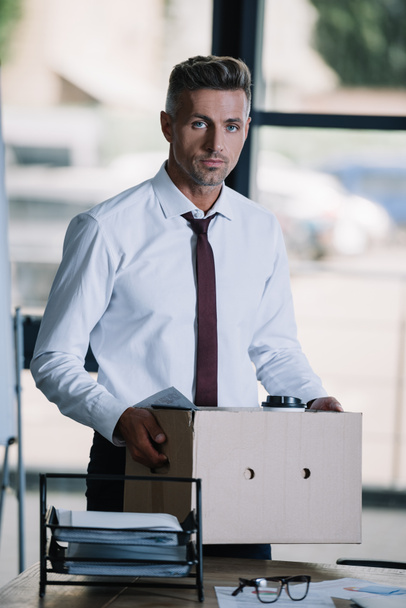 dismissed businessman holding box while standing near workplace and looking at camera  - Foto, Bild