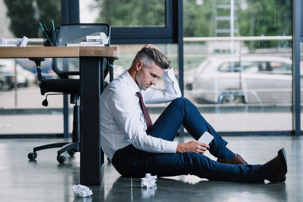 handsome businessman sitting on floor and holding smartphone near table in office  - Foto, Bild