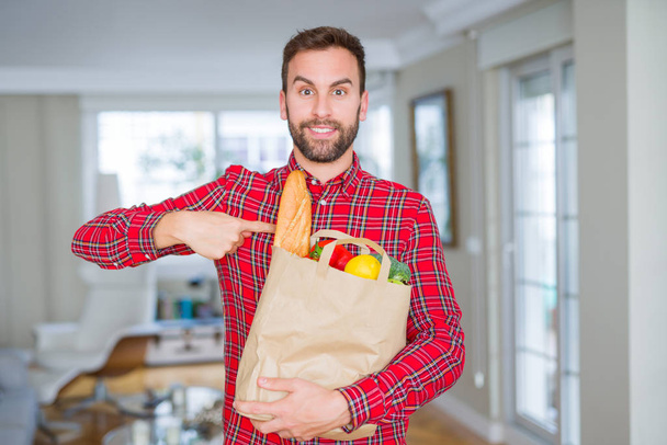 Hombre guapo sosteniendo bolsa de comestibles con la cara sorpresa señalando el dedo a sí mismo
 - Foto, Imagen
