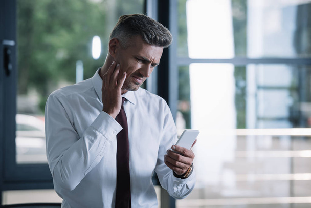 αναστατωμένος επιχειρηματίας σε κοστούμι κοιτάζοντας το smartphone στο γραφείο  - Φωτογραφία, εικόνα