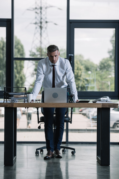 businessman standing near desk and laptop and looking at camera in office  - Foto, Imagem