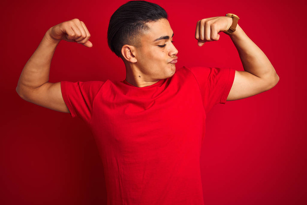 Jonge Braziliaanse man draagt t-shirt staande over geïsoleerde rode achtergrond toont Arms spieren glimlachend trots. Fitness concept. - Foto, afbeelding