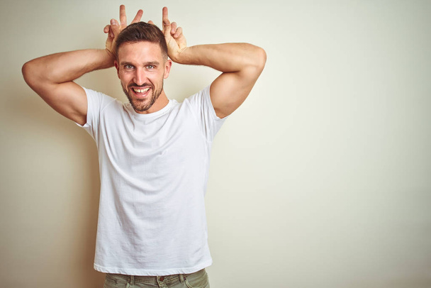 Όμορφος νεαρός φορώντας περιστασιακή λευκό t-shirt πάνω από το απομονωμένο φόντο που παριστάνουν τα αυτιά λαγουδάκι, αστεία και τρελή με δάχτυλα στο κεφάλι χαμογελώντας χαρούμενα - Φωτογραφία, εικόνα