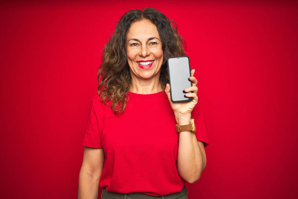 Старшая женщина средних лет показывает экран смартфона на красном изолированном фоне с счастливым лицом стоя и улыбаясь с уверенной улыбкой показывая зубы
 - Фото, изображение