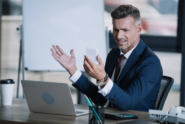 selective focus of upset man gesturing near laptop while holding smartphone  - Foto, Bild