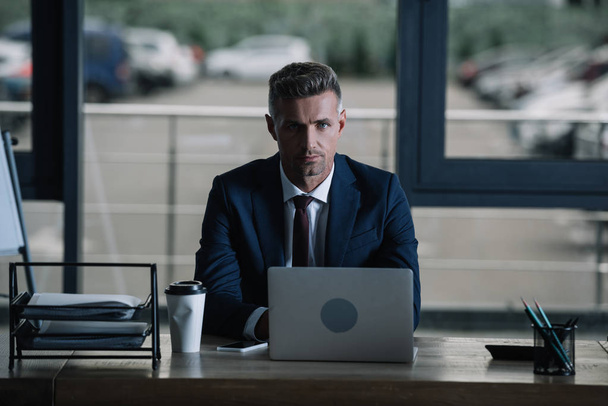 handsome businessman looking at camera near laptop and paper cup  - Foto, Imagem
