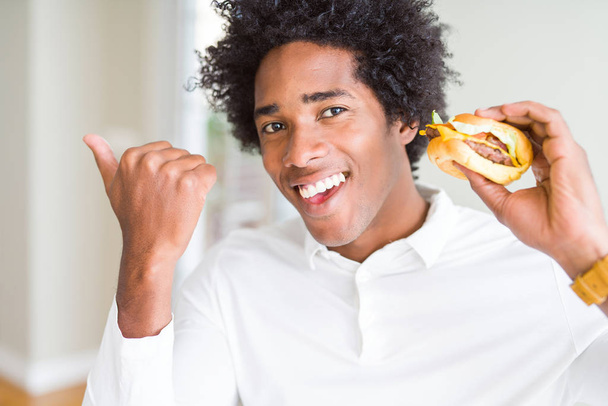 Afro-amerikai éhes ember étkezési hamburger ebédre mutatva és mutatja hüvelykujjával fel az oldalsó boldog arc mosolygós - Fotó, kép