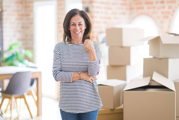 Middelbare leeftijd vrouw verhuizen naar een nieuw huis rond kartonnen dozen glimlachend met gelukkig gezicht kijken en wijzend naar de zijkant met duim omhoog. - Foto, afbeelding